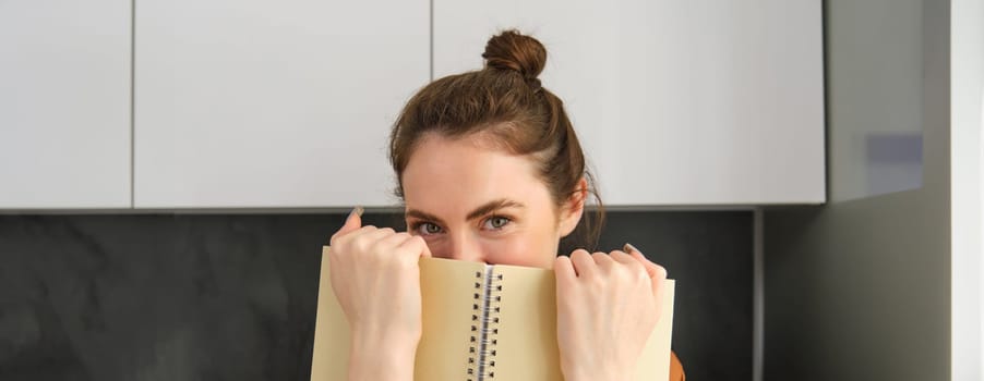 Cute romantic woman, hiding face behind notebook, smiling with eyes.
