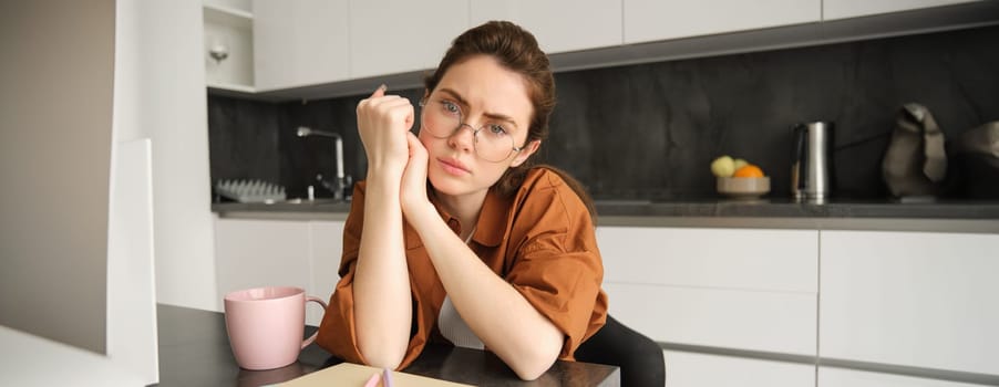 Portrait of sad and tired young woman in glasses, sitting in kitchen at home, looking upset and exhausted, working from home.
