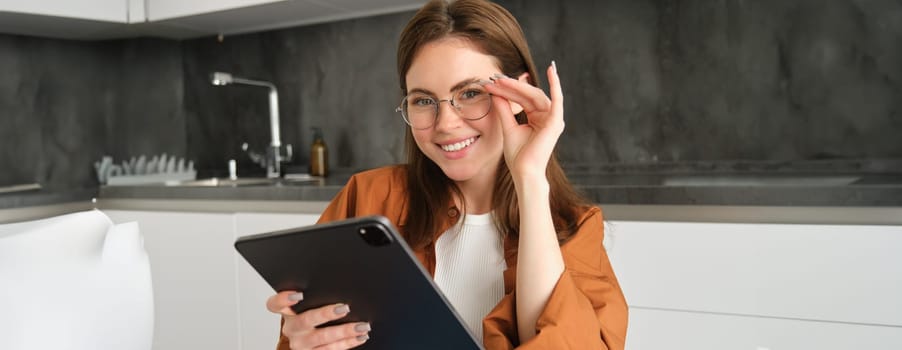 Portrait of confident, smiling young woman, student in glasses, sitting in kitchen, studying on remote, connects to online course on digital tablet.