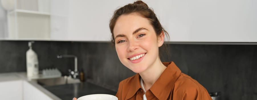 Close up portrait of beautiful brunette woman spends time, drinks delicious home brewed coffee, smiling at camera.