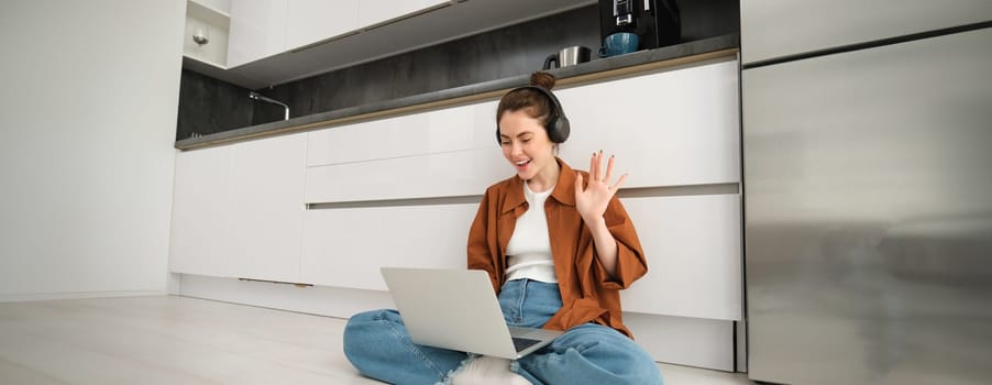 Portrait of happy woman in headphones, looking at laptop, waving hand at computer, chatting on video online, joins team meeting, calling friend on computer, sitting on floor.