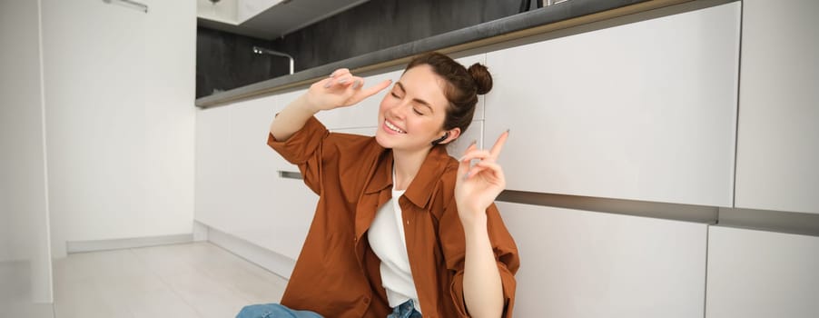 Portrait of young carefree woman sits on floor at home, dances while listens to music in wireless headphones, singing along favourite song.