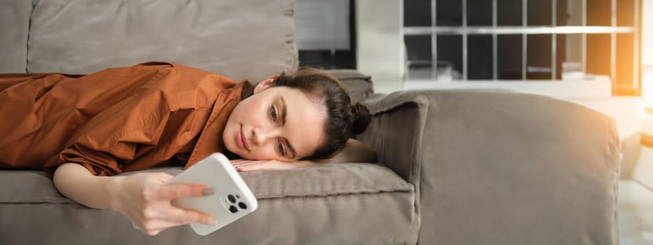 Close up portrait of smiling, beautiful young woman lying on sofa at home, checking messages on smartphone, resting on couch and looking at mobile phone.