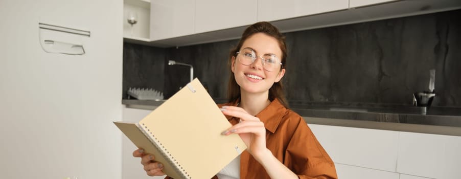 Close up portrait of beautiful young woman holding notebook, reading planner, sitting at home and studying, revising or doing homework.