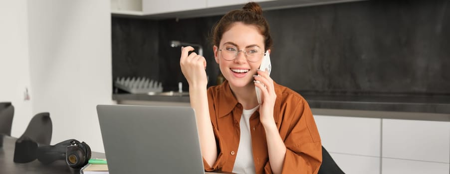 Portrait of beautiful smiling woman working from home, talking on mobile phone, calling client, self-employed businesswoman sets up workplace in kitchen, using laptop and smartphone.