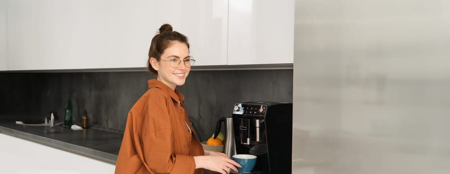 Portrait of young woman making herself coffee, standing near machine and smiling, holding mug for cappuccino, looking happy at camera.