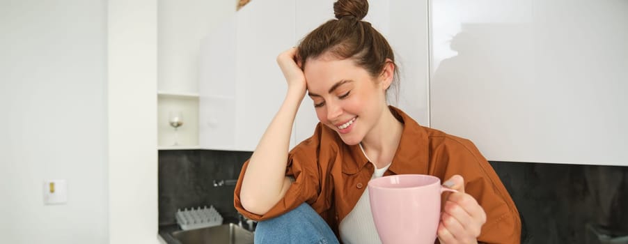 Portrait of beautiful young woman enjoying her morning cup of aromatic coffee, drinking tea while sitting in kitchen and contemplating sunny day.