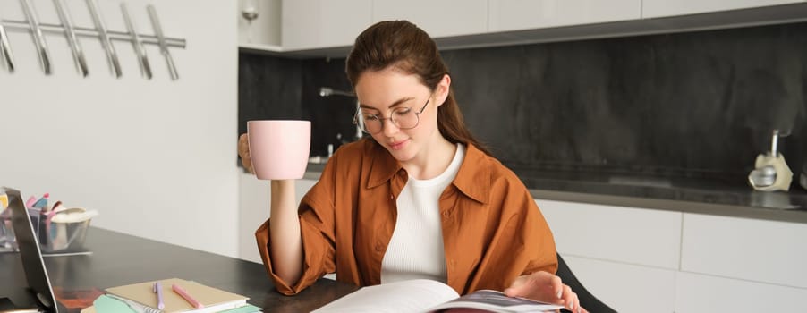 Distance learning. Young woman at home, student studying at home and drinking tea, reading her work book, revising for exam.