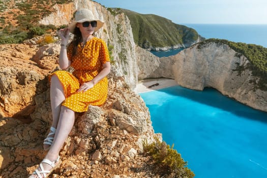 One beautiful young Caucasian brunette girl in a yellow dress with polka dots, sunglasses and a straw hat sits on the edge of a cliff against the backdrop of a deserted beach with a sunken ship on the island on a summer sunny day, side view from the mountain close-up.