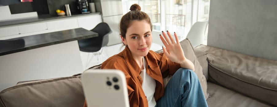 Happy woman is sitting at home with smartphone, online chatting, waving hand at mobile phone, saying hello, talking to friends using application, sitting on sofa at home.
