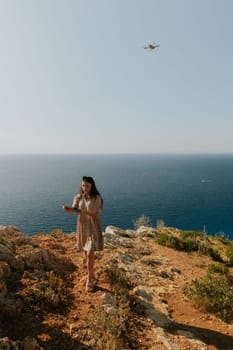 One young beautiful Caucasian brunette girl films herself on a drone flying behind her, holding a control panel in her hands, standing on a mountain top against the backdrop of a blurred sea, close-up side view.