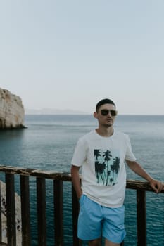 One handsome handsome brunette guy in sunny clothes stands on a wooden terrace against the backdrop of the sea and rocks on a sunny summer evening, close-up side view.