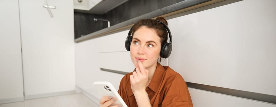 Close up of young woman sits on floor at home, listens music in headphones, holds smartphone, looks thoughtful, orders online on mobile phone app.