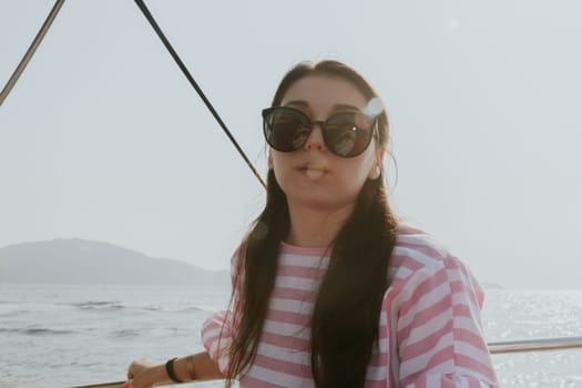 Portrait of one beautiful Caucasian brunette woman in sunglasses and a pink striped dress sitting in a boat and sailing in the sea on a sunny summer day, close-up side view.