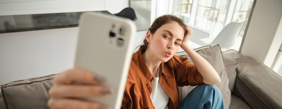 Portrait of cute young woman, modern girl taking selfies on smartphone app, posing on couch, extends her white mobile phone to take picture, pucker lips for photo.