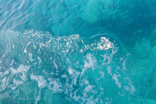 Beautiful view of blue colored water with a white foamy wave in the center on a sunny summer day, close up view from above.