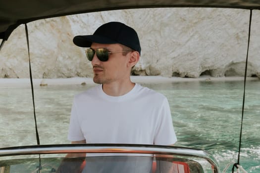 One young Caucasian guy in a cap and sunglasses looks to the left and sails alone on the sea on a boat, steering the helm on a sunny summer day, side view close-up.