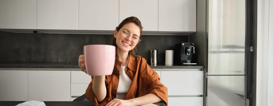 Portrait of happy, beautiful brunette woman in kitchen, student taking lunch break for cup of tea, raising mug, giving you coffee.