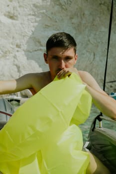 Portrait of one young handsome Caucasian brunette guy inflates a light green lifebuoy with his mouth while sitting in a boat against the backdrop of blurred rocks on a sunny summer day, side view close-up.
