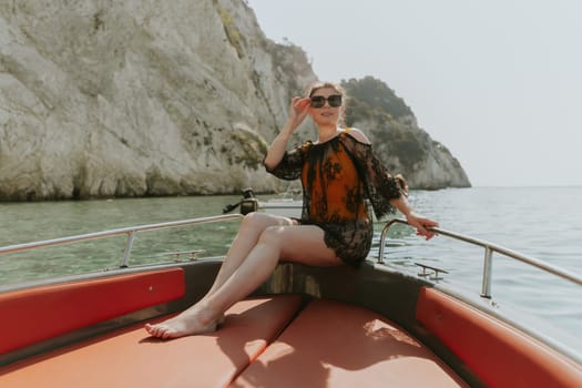 Portrait of one beautiful Caucasian brunette in sunglasses and a black lacy cape looking at the camera and sitting on the stern of a boat while sailing on the sea against the backdrop of a blurred rocky beach on a sunny summer day, close-up side view.