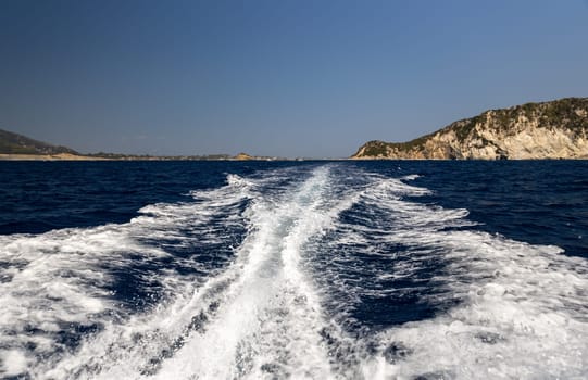 A beautiful view of foamy waves cutting from the motor of a boat in the blue sea on a sunny summer day against the backdrop of a blurred rocky island, close-up side view.