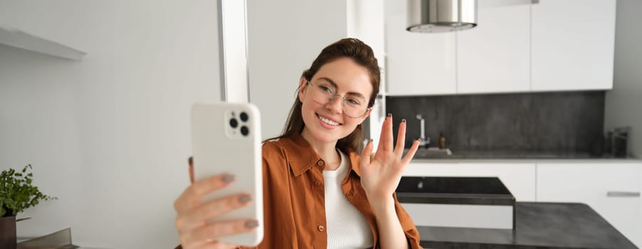 Portrait of stylish beautiful woman at home, video chats on smartphone, connects to online meeting from her living room, lifestyle social media blogger records a video on mobile phone.