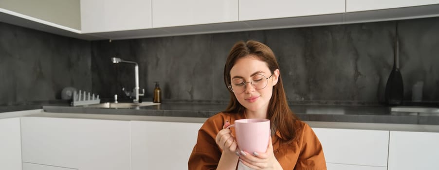 Relaxing day at home. Smiling young woman in glasses, enjoying fresh brewed cup of coffee, holding mug and sitting in kitchen, resting on peaceful autumn day at home.