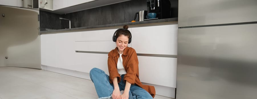 Portrait of young beautiful woman in wireless headphones, sitting on kitchen floor, vibing with favourite music in earphones.