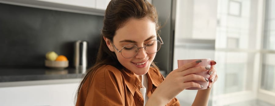 Close up portrait of woman in glasses, smiling and laughing, holding cup of tea, drinking coffee at home, resting with mug of fresh delicious coffee.