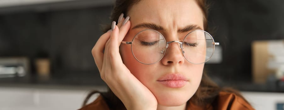 Portrait of working woman at home, student has headache, touches head and has painful migraine, sits in kitchen in glasses.