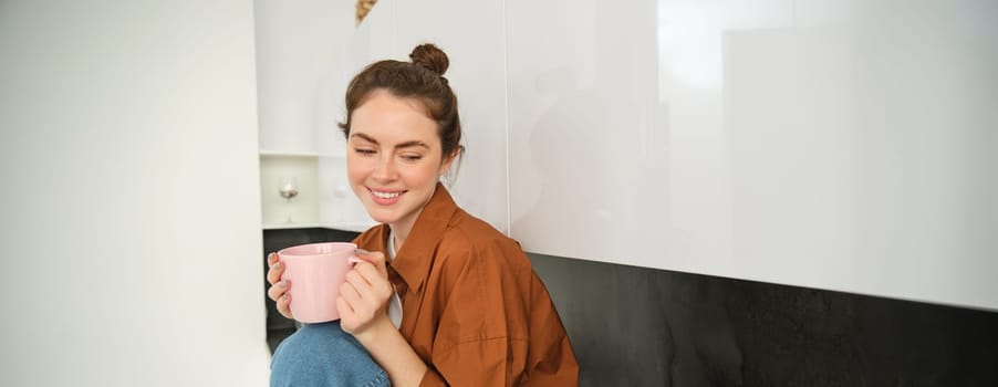 Portrait of young woman with cup of coffee, sits in kitchen and drinks aromatic drink at home, holds tea mug.