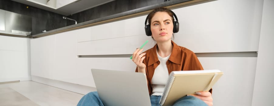 Portrait of young woman, modern student sits at home on floor and doing homework on laptop, listens music in headphones while making assignment or project for college.