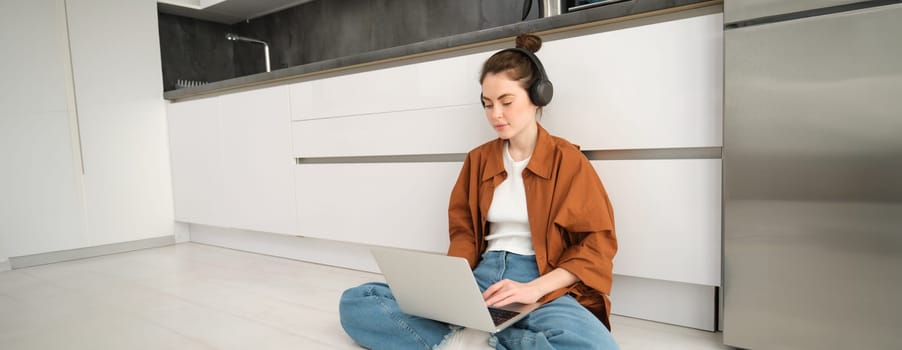 Young woman freelancer, working from home, student sitting with laptop on kitchen floor, wearing headphones, typing on computer.
