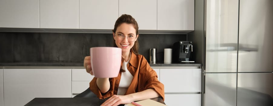 Stylish young woman, student studying from home, sitting in kitchen with homework, giving you cup of coffee, raising pink mug, smiling at camera.