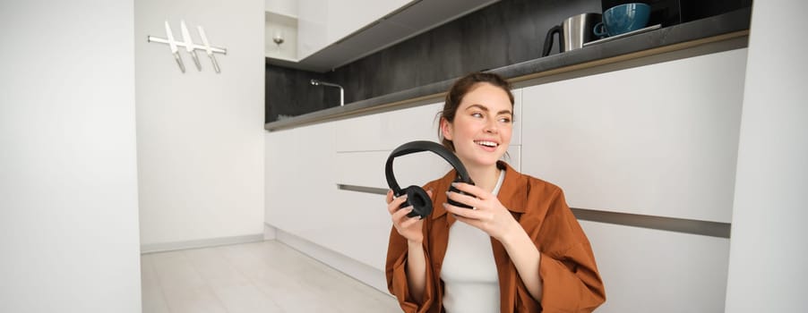 Carefree modern woman sits on floor at home, smiles and looks aside, puts on wireless headphones to listen to music or podcast.