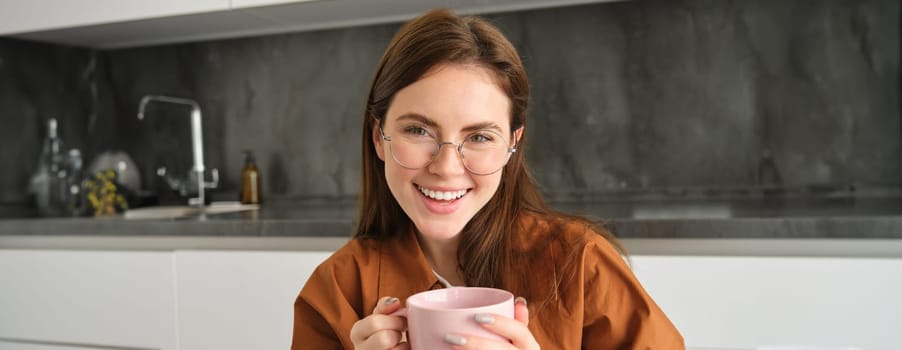 Portrait of young brunette woman in kitchen, resting indoors, smiling and drinking coffee, enjoys tasty hot tea.