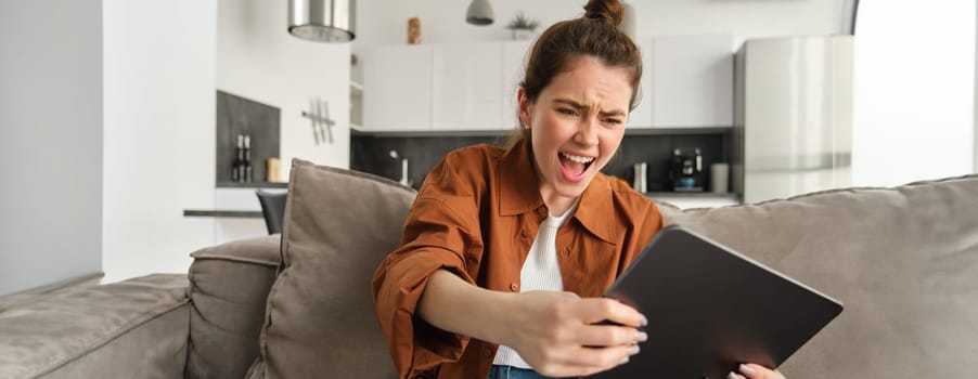 Portrait of young woman playing video games on digital tablet, looking excited, tilting gadget and looking at screen, sitting on couch in living room.