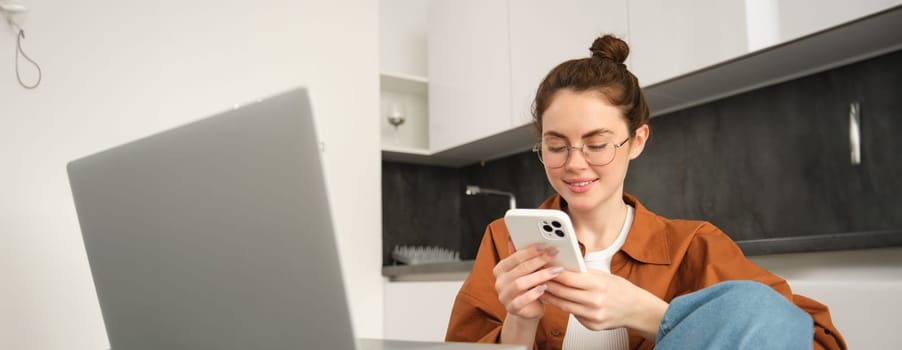 Portrait of smiling young woman in glasses, sitting with laptop and smartphone in kitchen, working from home, student studying, doing homework.