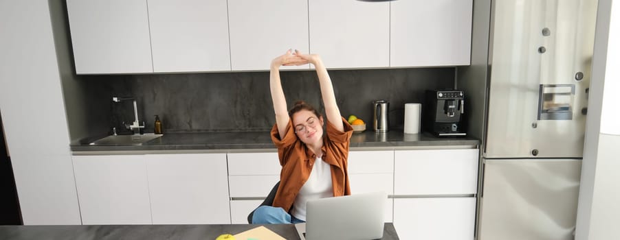 Portrait of smiling, pleased woman in glasses, sitting in kitchen, finish working on project, using laptop, stretching hands with satisfied face expression.