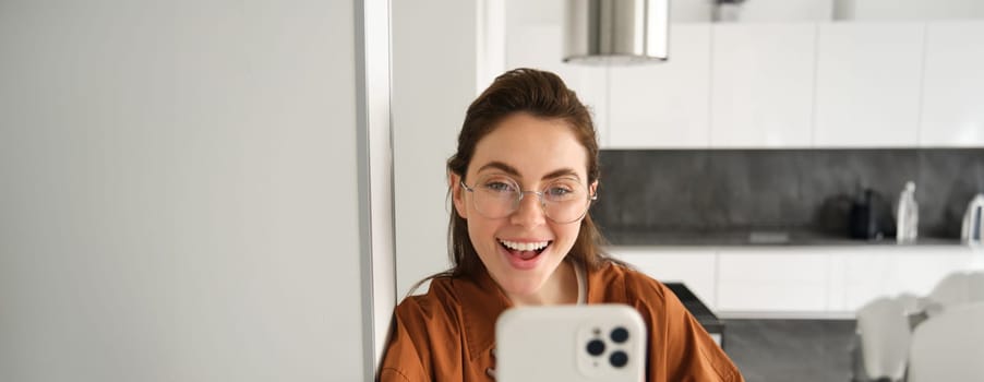 Close up portrait of happy girl takes selfie on smartphone, posing with mobile phone, looking excited and amazed, smiling at telephone camera, using social media app.