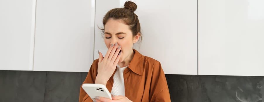 Young stylish woman at home, looking at mobile screen, yawning while reading a message.