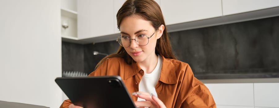 Portrait of young student, woman studying at home, working remotely, setup workplace in her kitchen, sitting on chair with digital tablet, reading in glasses.