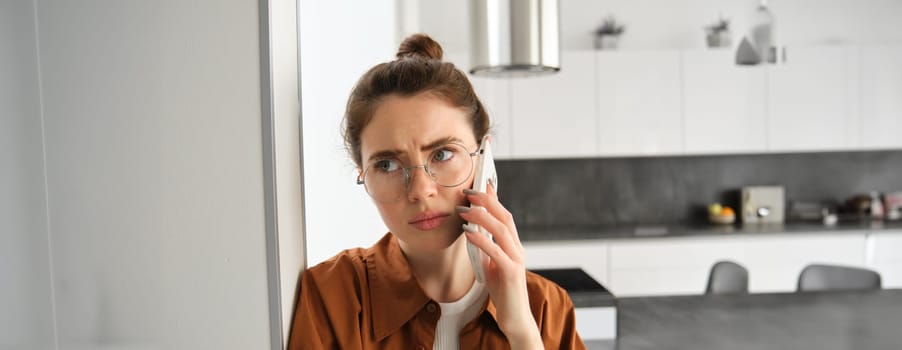 Portrait of woman with disappointed face, standing at home, answers phone call with concerned face, has difficult telephone conversation.