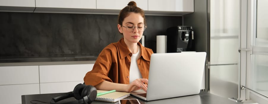 Work and life balance concept. Young woman in glasses, working from home, typing on laptop, student doing homework on computer, sitting in kitchen, freelancing.