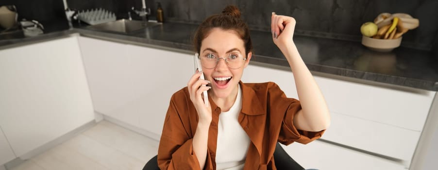 Portrait of woman receiving great news over the phone. Girl talking on mobile telephone and celebrating, laughing and making fist pump, dancing on her chair in kitchen.