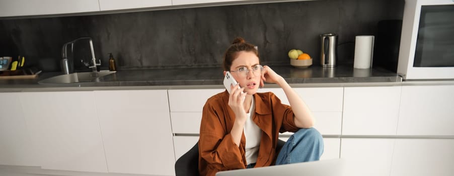 Portrait of woman working from home in glasses, sitting with confused face after answering a phone call, has laptop on table.