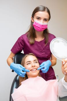 Young woman smiling while looking at mirror while doctor demonstrating the results of work in dental clinic