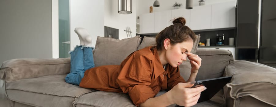 Portrait of woman resting on couch at home, rubs her eyes after reading too long on digital tablet, feeling tired, spending time in living room on sofa.