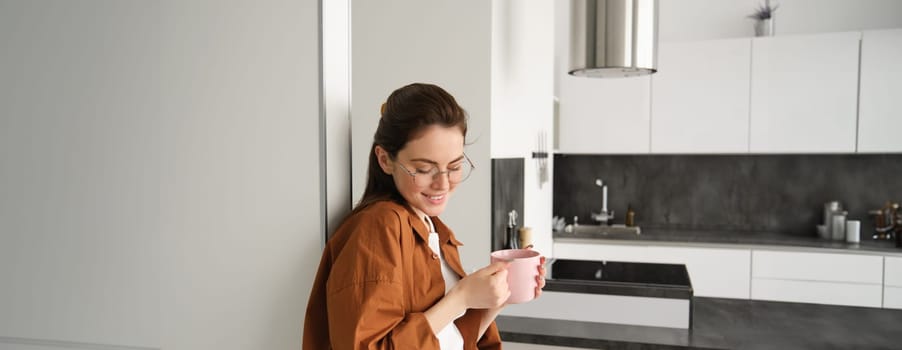 Portrait of cosy, beautiful and relaxed young woman at home, standing and leaning on wall with cup of coffee, drinking tea with thoughtful face expression.
