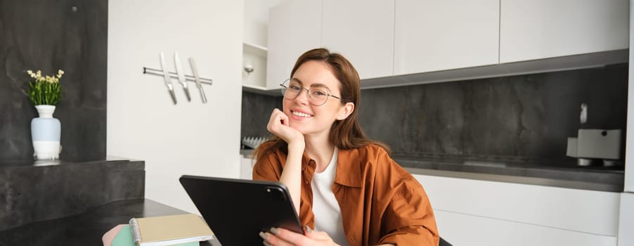 E-learning and remote workplace concept. Young woman in glasses, student studying at home in kitchen, reading on digital tablet, looking at her project, connects to online meeting.